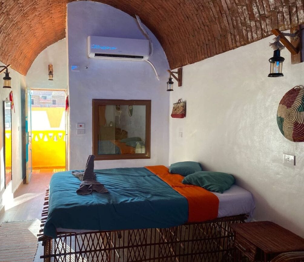 A room with a bed, a wooden table, a wall-mounted air conditioner, and a window with a view of the outside.