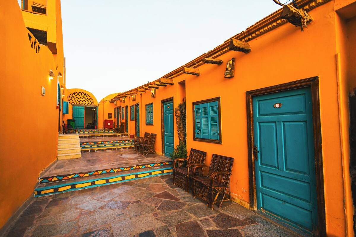 A narrow alleyway with a stone floor, wooden chairs, and a blue door.