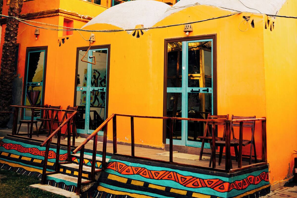A yellow building with a balcony, a staircase, and palm trees in the background.