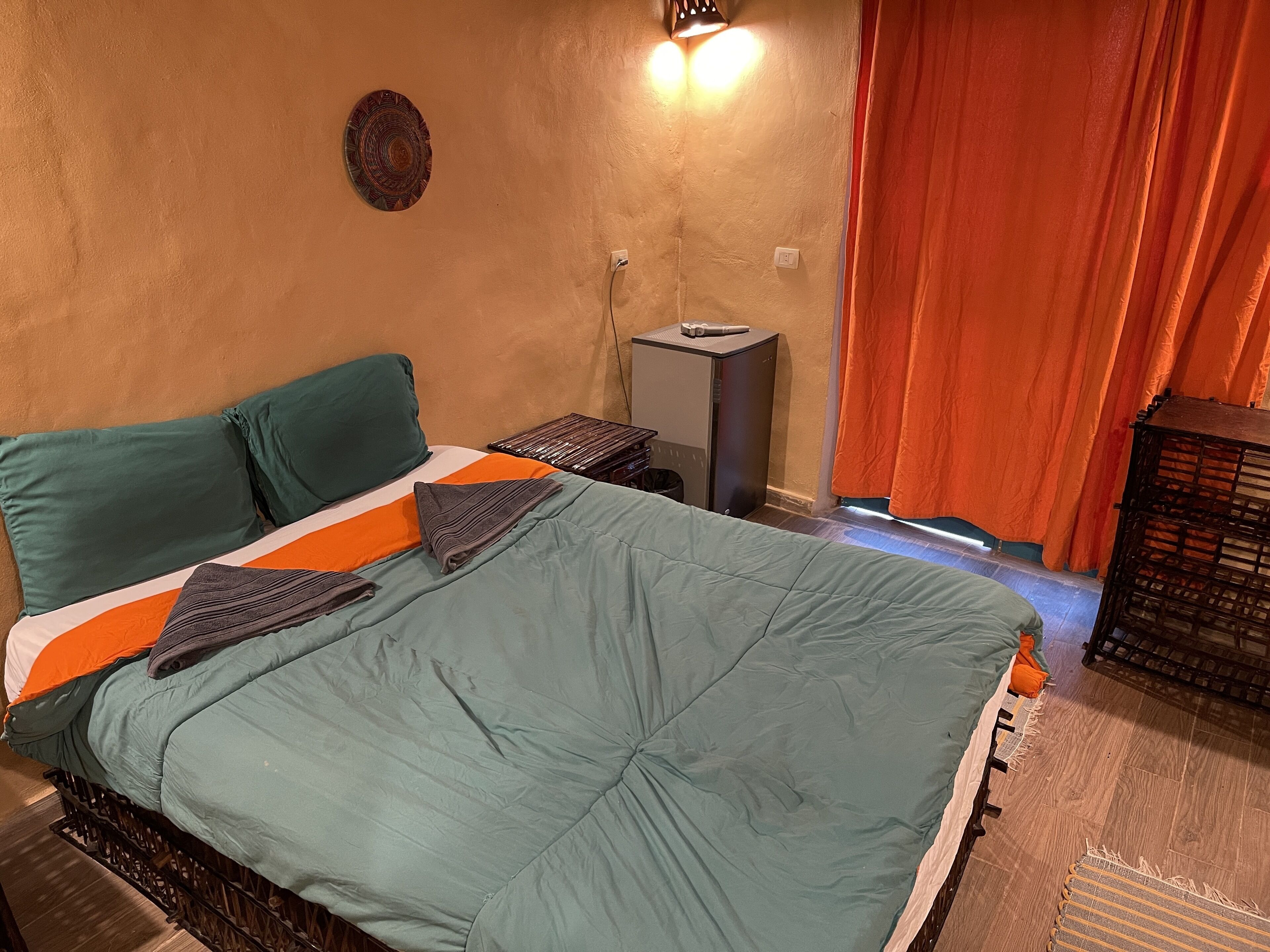 A neatly made bed with a teal comforter and pillows, a small bedside table with a lamp, and a window with orange curtains.