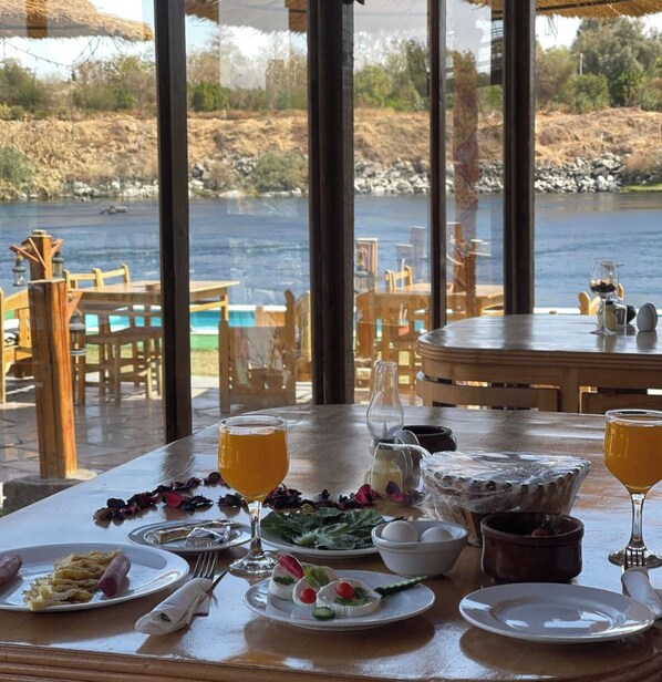 A dining area with a view of a lake, featuring a table set with food and drinks, and a sign indicating a free airport shuttle service. - Fenti Nubian Resort (Aswan)