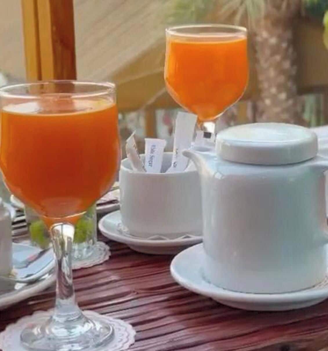 Een tafel met twee glazen sinaasappelsap, een witte theepot en een suikerpot met zakjes.