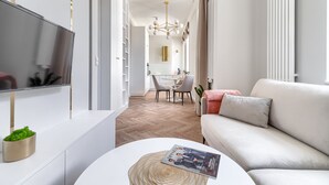 Een moderne woonkamer met een bank, een flatscreen-tv gemonteerd op een witte kast, en een eethoek op de achtergrond.