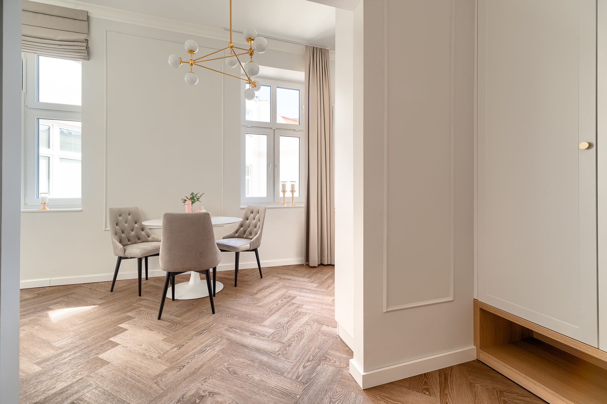 A modern dining area with a wooden herringbone floor, two upholstered chairs, a round table, and a chandelier.