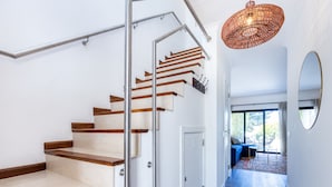Un interior moderno con una escalera de madera, un colgante de luz y vistas a un balcón con mobiliario exterior.