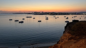 Escena costera con varios veleros anclados en aguas tranquilas, un horizonte urbano lejano y un litoral rocoso.