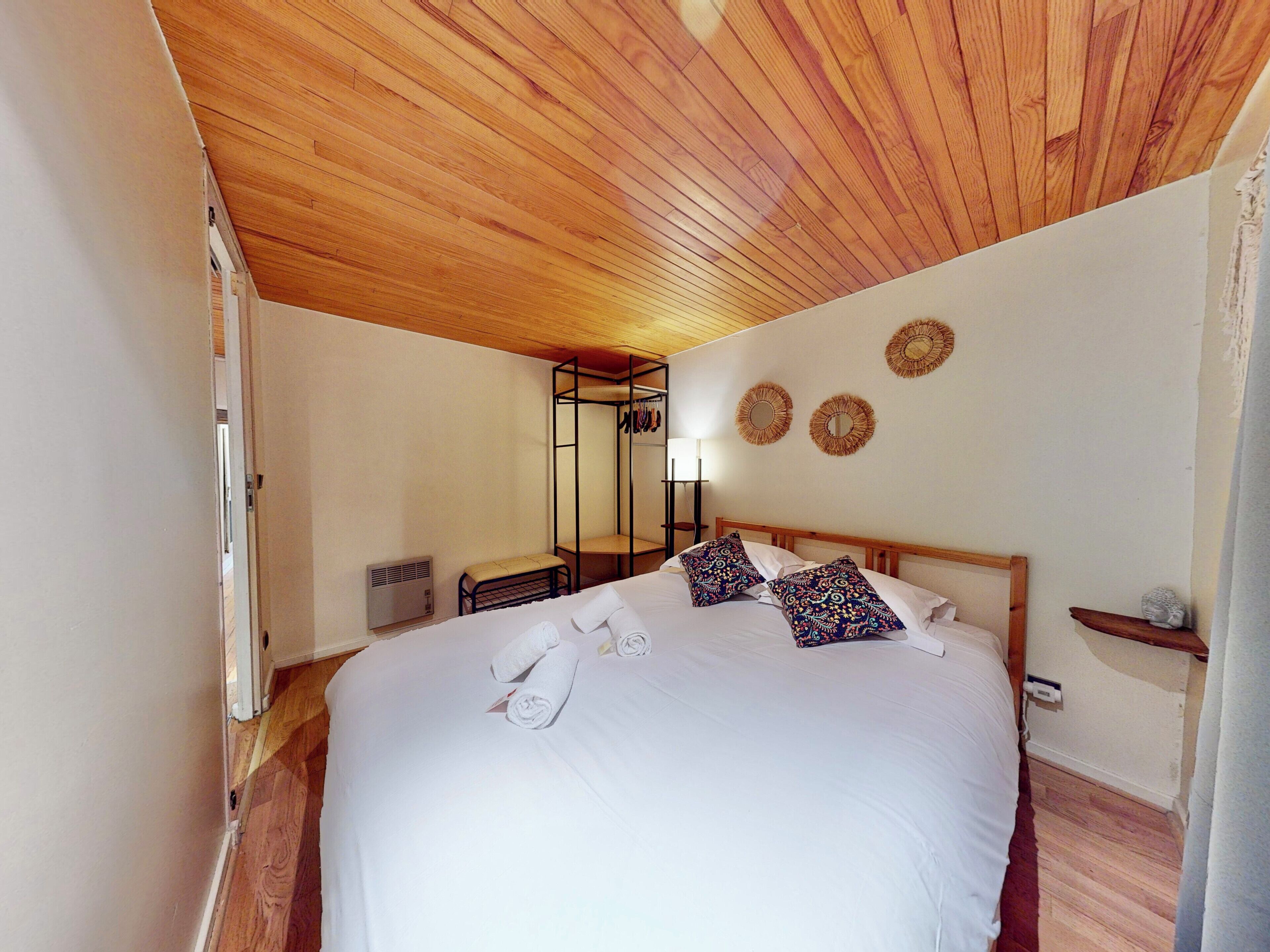 A bedroom with a wooden ceiling, a bed with white linens and patterned pillows, a bedside table, a mirror, and wall decorations.