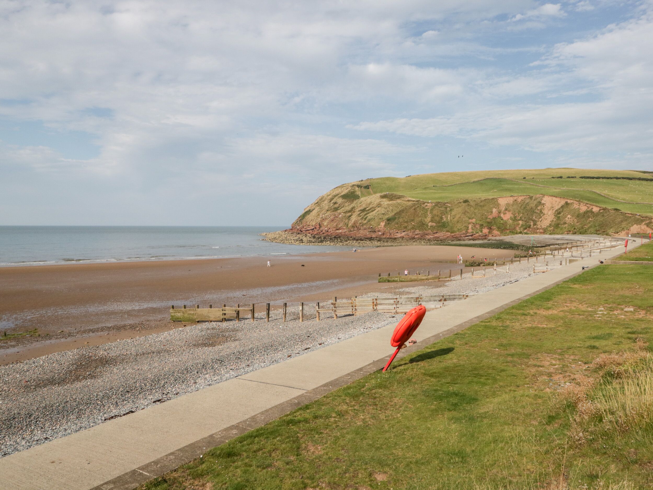 A coastal beach with a concrete walkway, a grassy area, and a red buoy.