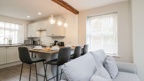 Cozinha moderna com armários brancos, uma ilha central com banquetas, iluminação pendente e uma grande janela com persianas.