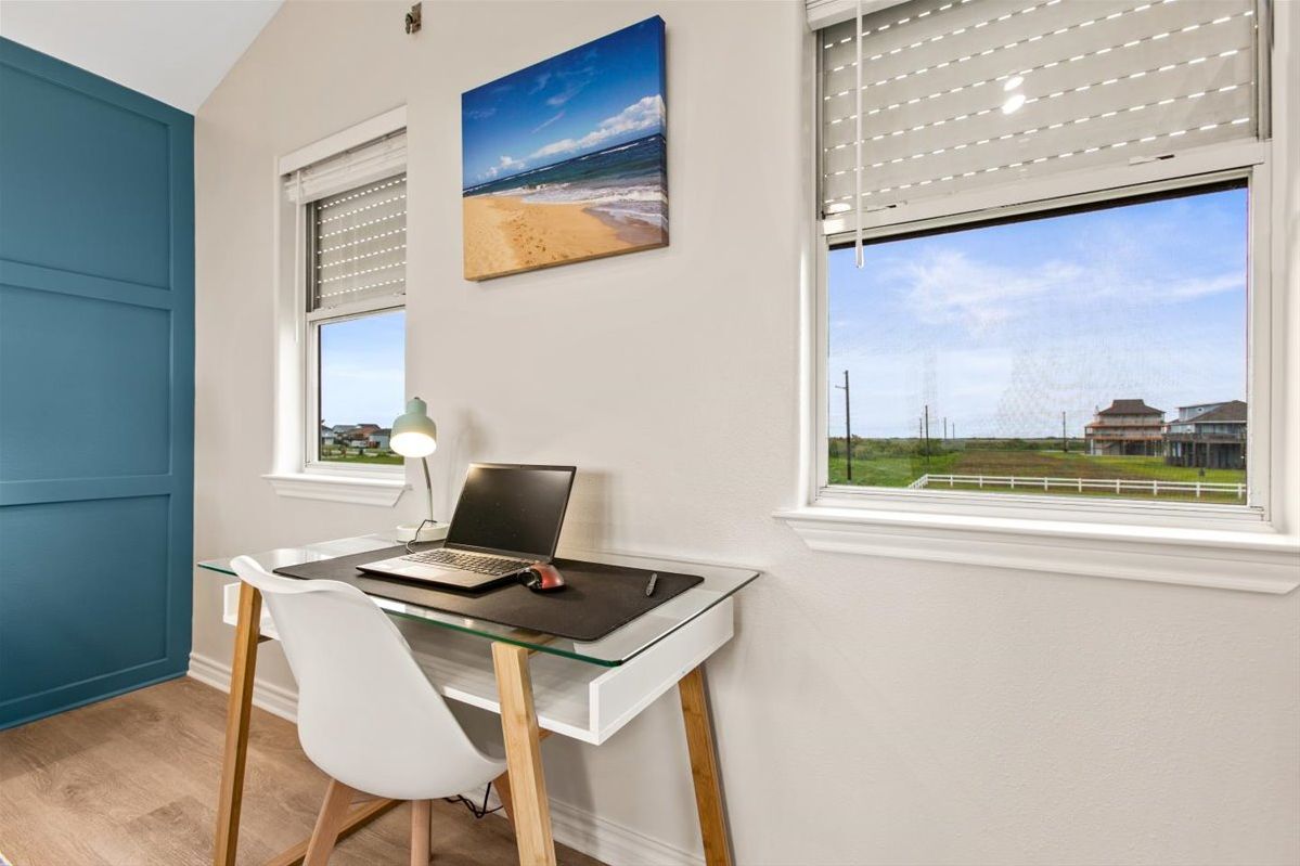 Une pièce avec un bureau en verre, un ordinateur portable, une chaise blanche et un tableau représentant une plage accroché au mur.