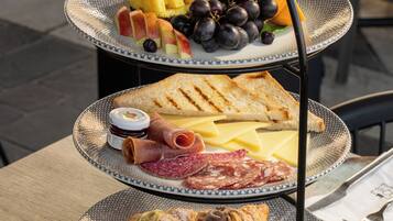 A three-tiered outdoor dining table with a variety of food items, including fruits, cheeses, meats, and pastries, set against a backdrop of a fire.