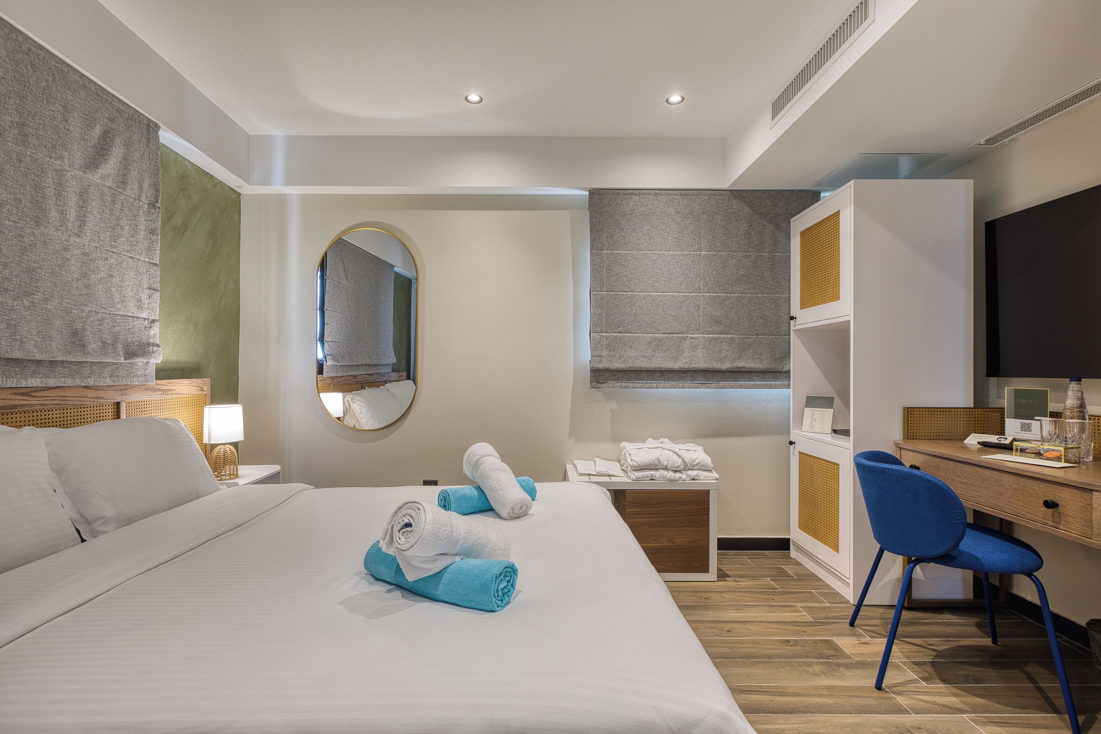 A modern hotel room with a large bed, a desk with a computer, a chair, and a wardrobe.