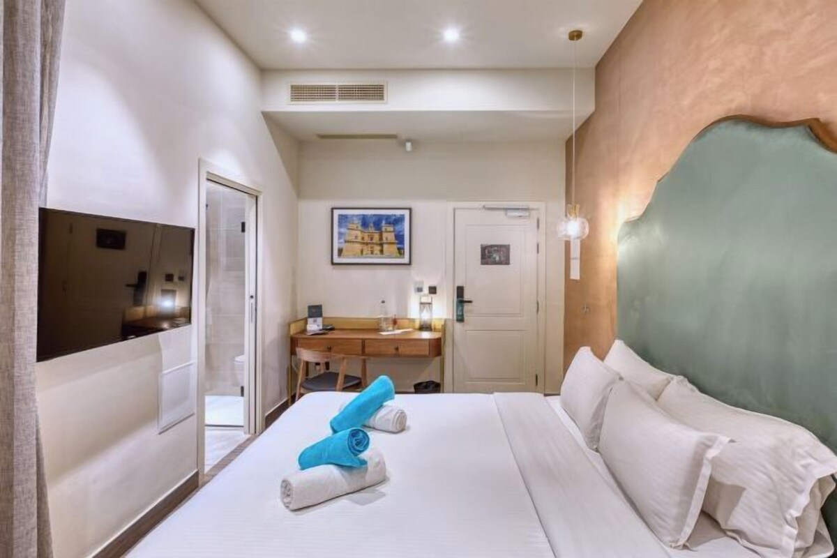 Hotelkamer met een bed, een bureau, een stoel en een badkamer zichtbaar achter een glazen wand.