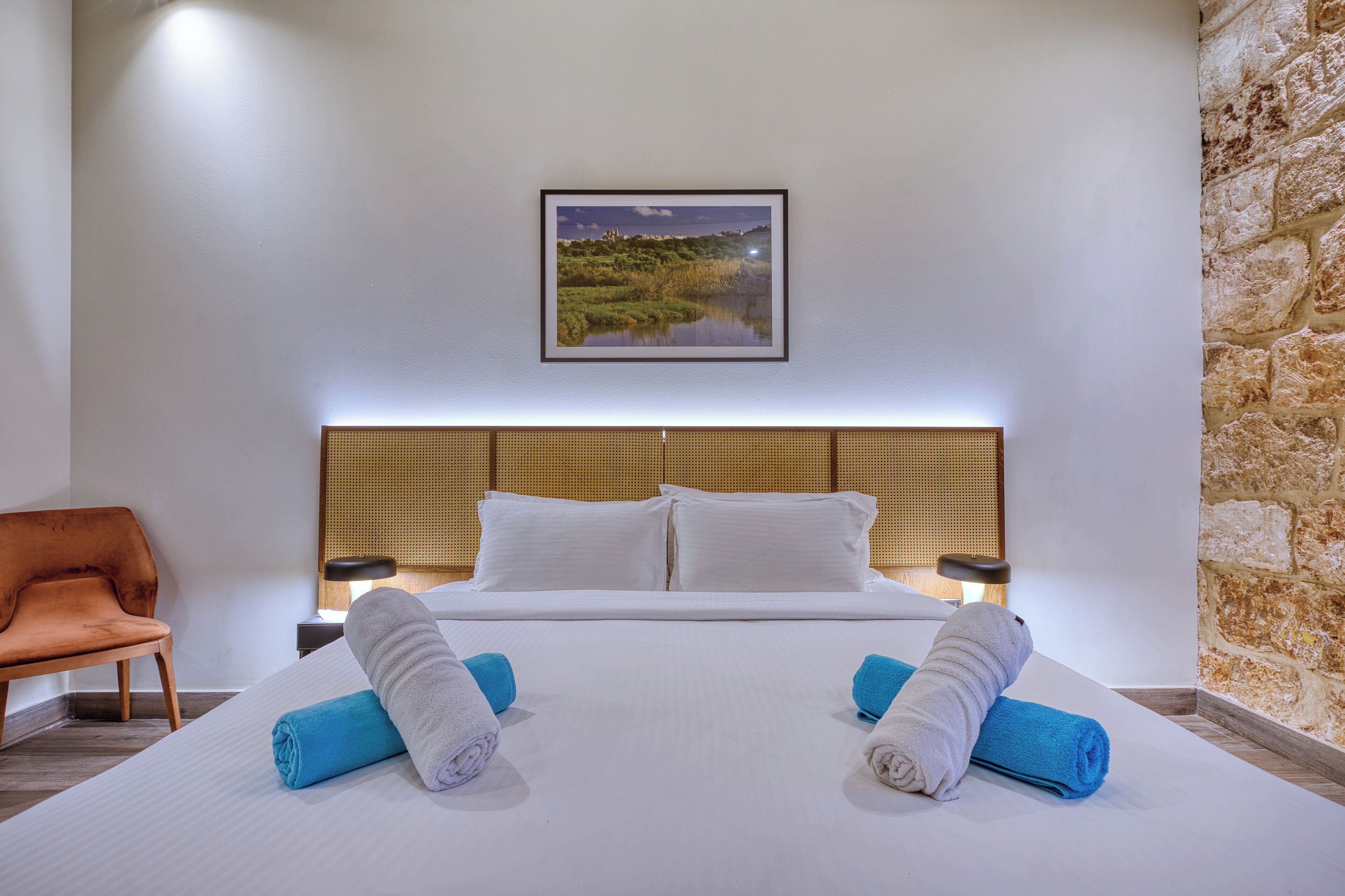Habitación de hotel con una cama grande, dos almohadas, una mesita de noche con una lámpara, un cuadro enmarcado en la pared y una pared decorativa de piedra.