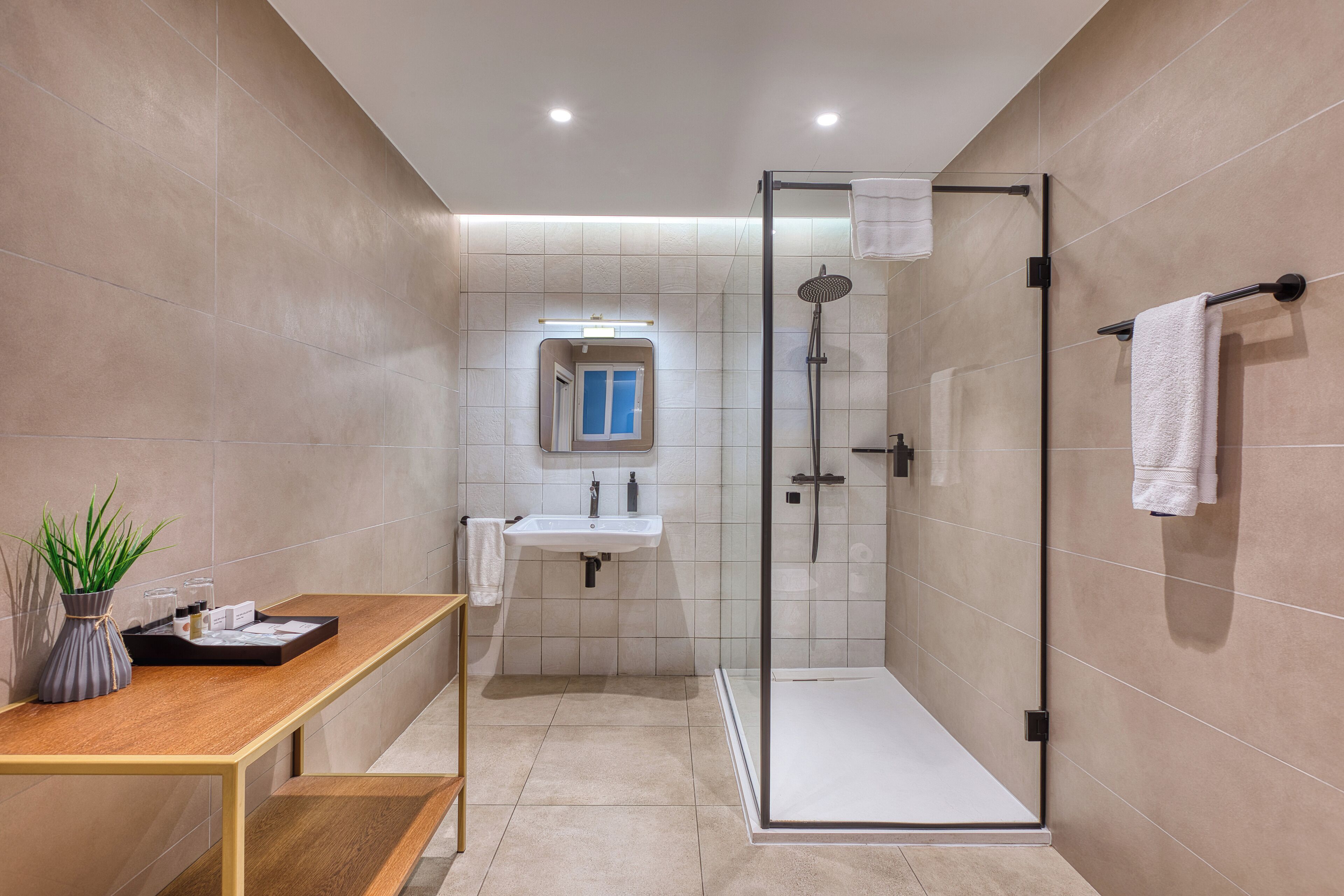 Un baño moderno con ducha de vidrio, un lavamanos con espejo y una banca con artículos de aseo.