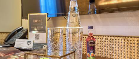 Zona de recepción de un hotel con un expositor de vidrio que contiene un macaron, una bolsita pequeña y un trozo de chocolate. Hay tres botellas de vidrio transparente, una botella de licor Zepp's Baxtra y un teléfono negro sobre el mostrador. Se ve un menú de servicio a la habitación al fondo.