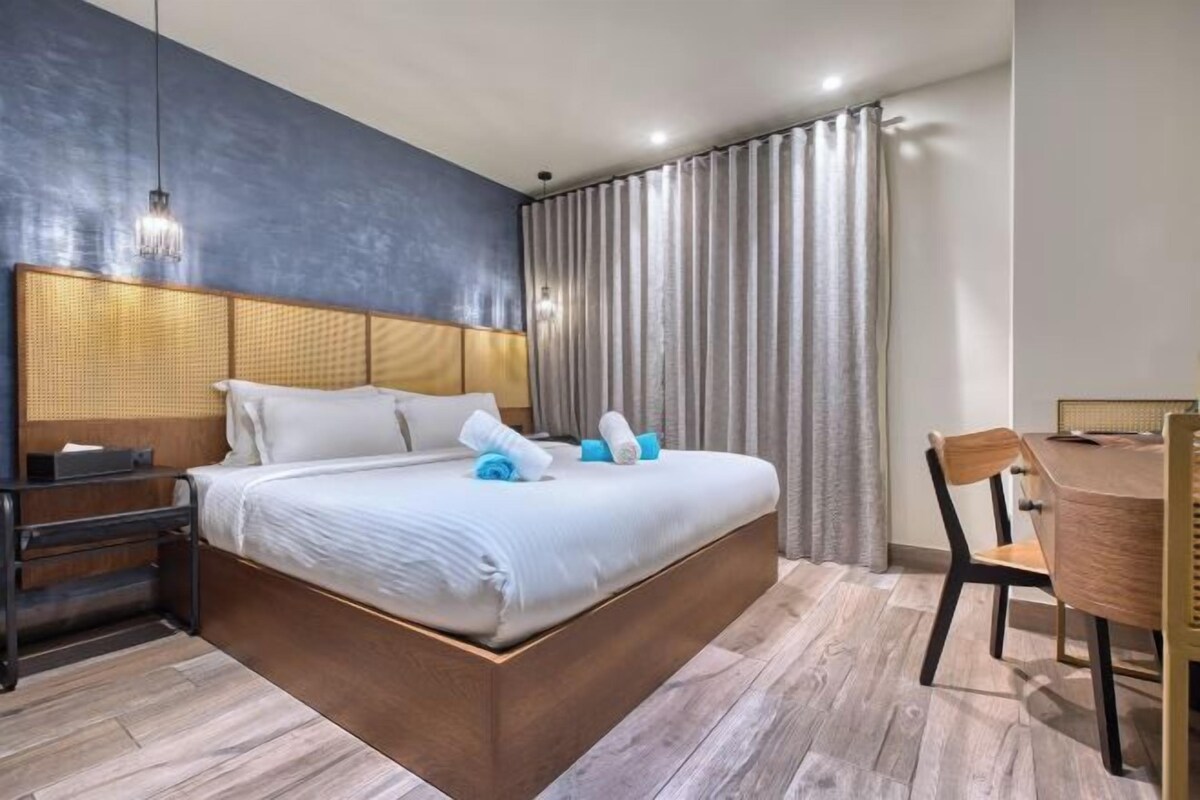 A modern hotel room with a large bed, a wooden headboard, a desk, and a chair.