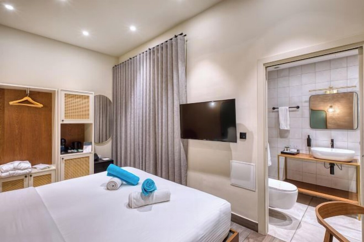 Habitación de hotel con cama, televisión, baño con lavamanos e inodoro, y armario.