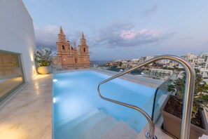 Piscina infinita no terraço com guarda-corrimão metálico, plantas em vasos e vista para uma igreja histórica e paisagem urbana.