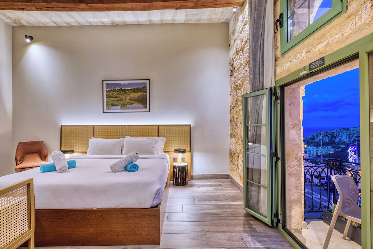 Habitación de hotel con cama, una silla, mesita de noche y balcón con vistas a la ciudad.
