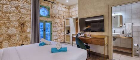 Un dormitorio con pared de piedra, una cama con ropa de cama blanca, un escritorio con una silla, un televisor y un baño con espejo y lavamanos.
