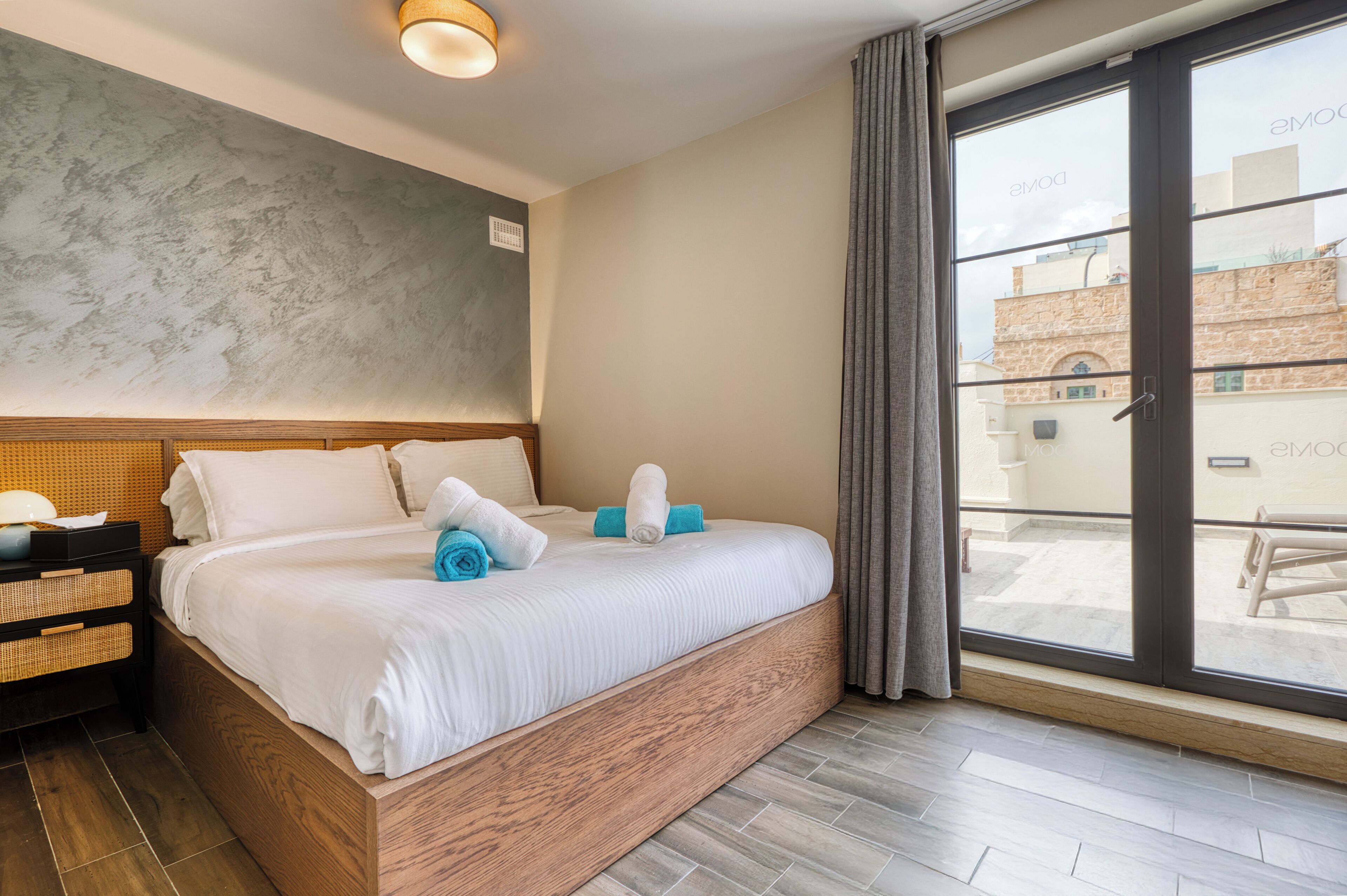 A modern hotel room with a large bed, a wooden headboard, a nightstand, and a view of the city through a sliding glass door.