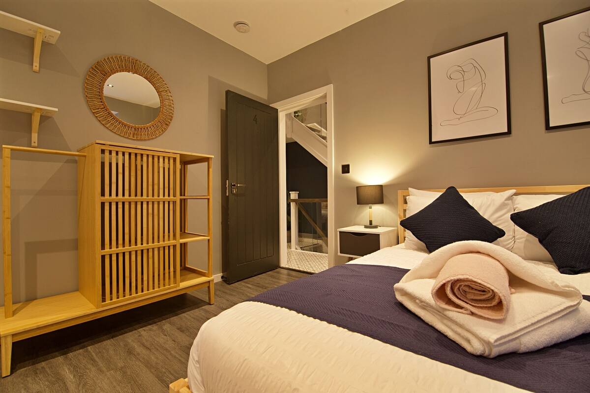 A bedroom with a bed, a wooden wardrobe, a mirror, and two framed artworks on the wall.