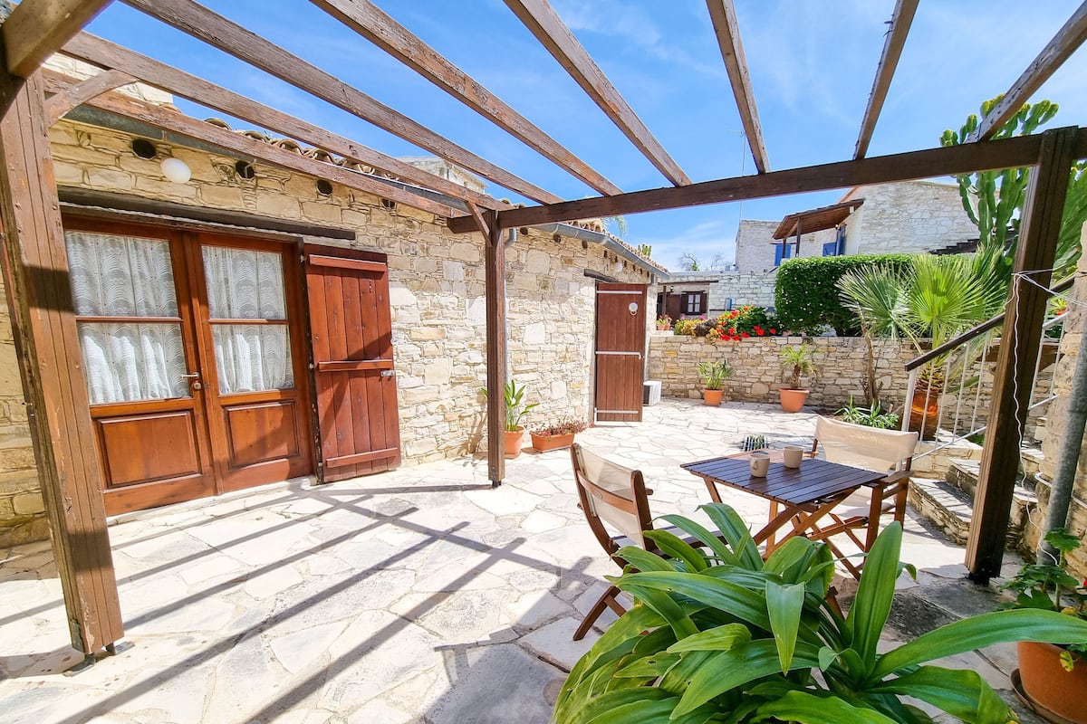 A stone building with wooden doors and a pergola. There is a table and chairs outside.