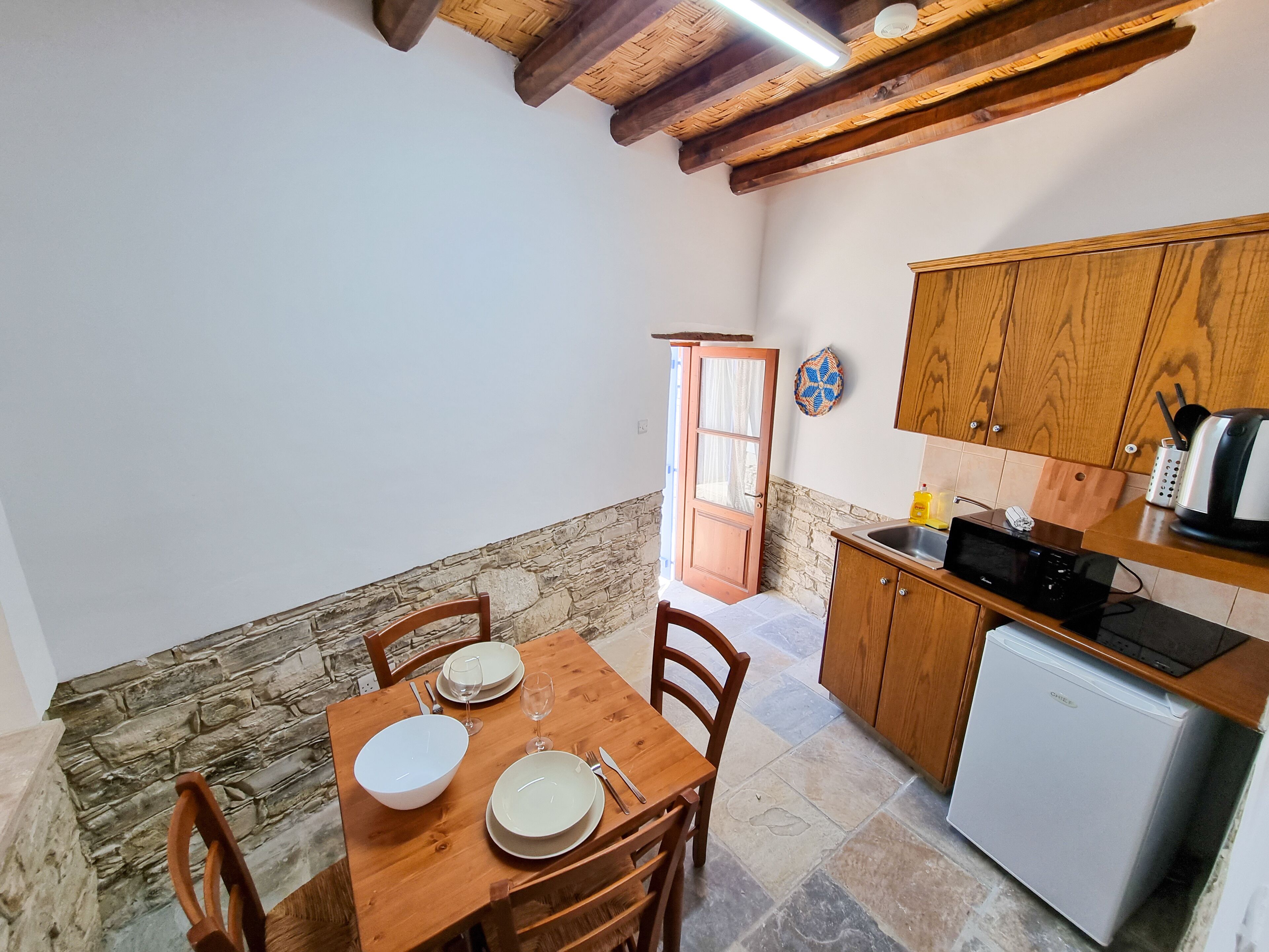 A cozy kitchen with a dining table set for two, wooden cabinets, a small refrigerator, and a stone wall.