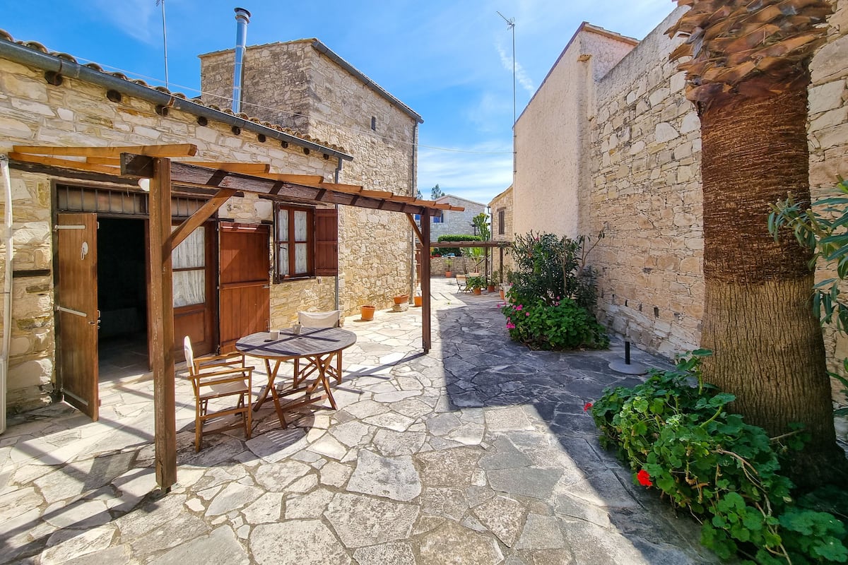 A stone courtyard with a wooden pergola, a table with chairs, and potted plants.