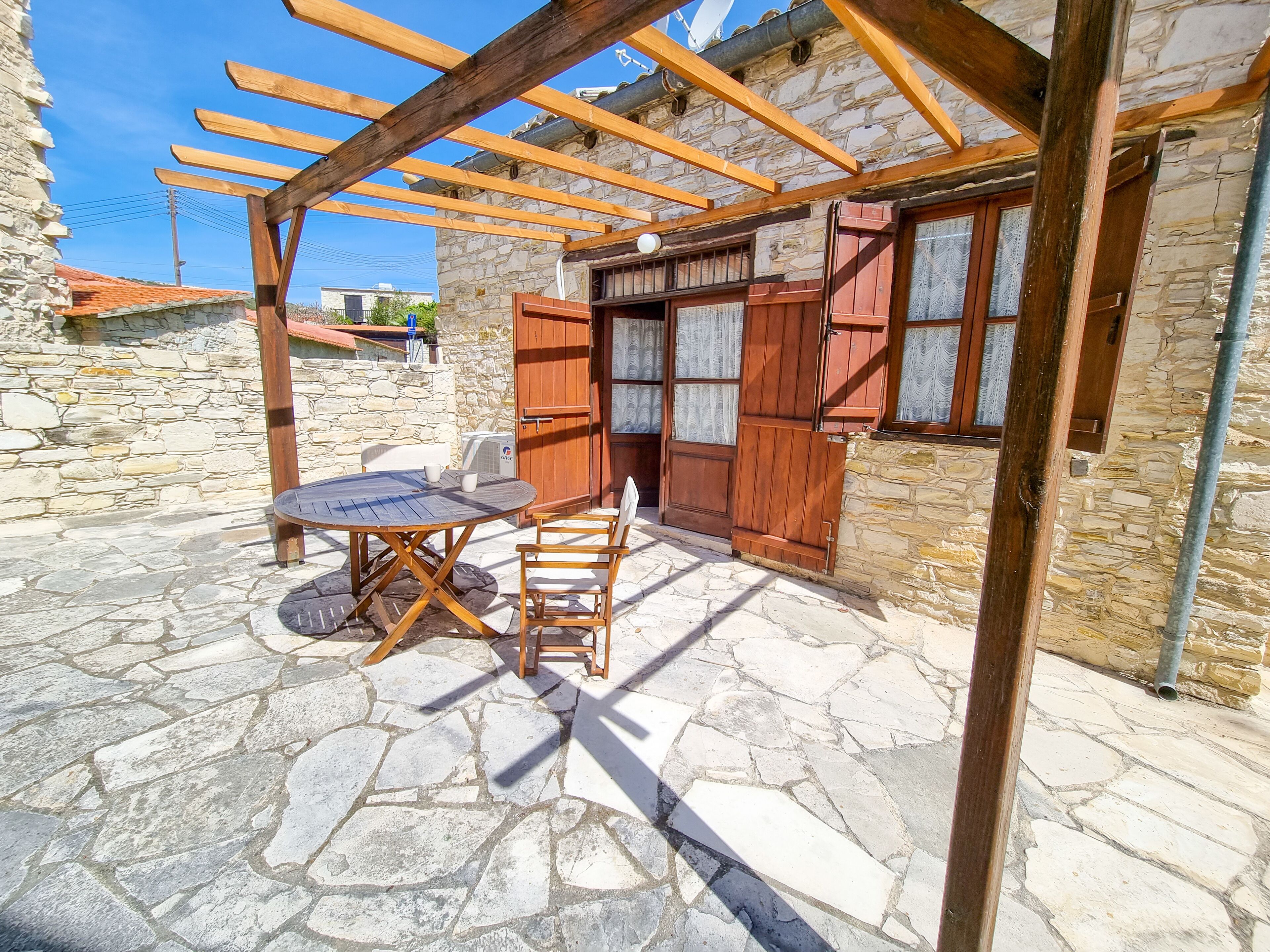 Une terrasse dallée de pierres, avec une table et des chaises en bois, une pergola et un mur en pierre.