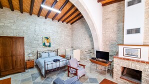 Una habitación rústica con pared de piedra, vigas de madera en el techo, una cama, un televisor, una chimenea y un cuadro.
