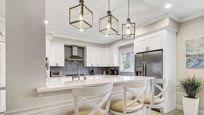 A modern kitchen with white cabinetry, a central island with bar stools, and pendant lighting. - Massive Luxury Beach Home Private Pool Hilton Head (Hilton Head Island)