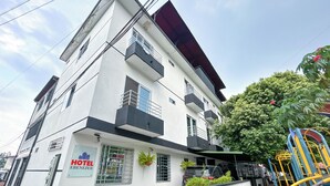 Un hotel moderno con un letrero que dice "HOTEL EBENIZER". Tiene fachada blanca con detalles negros, varios balcones y un estacionamiento visible al fondo.