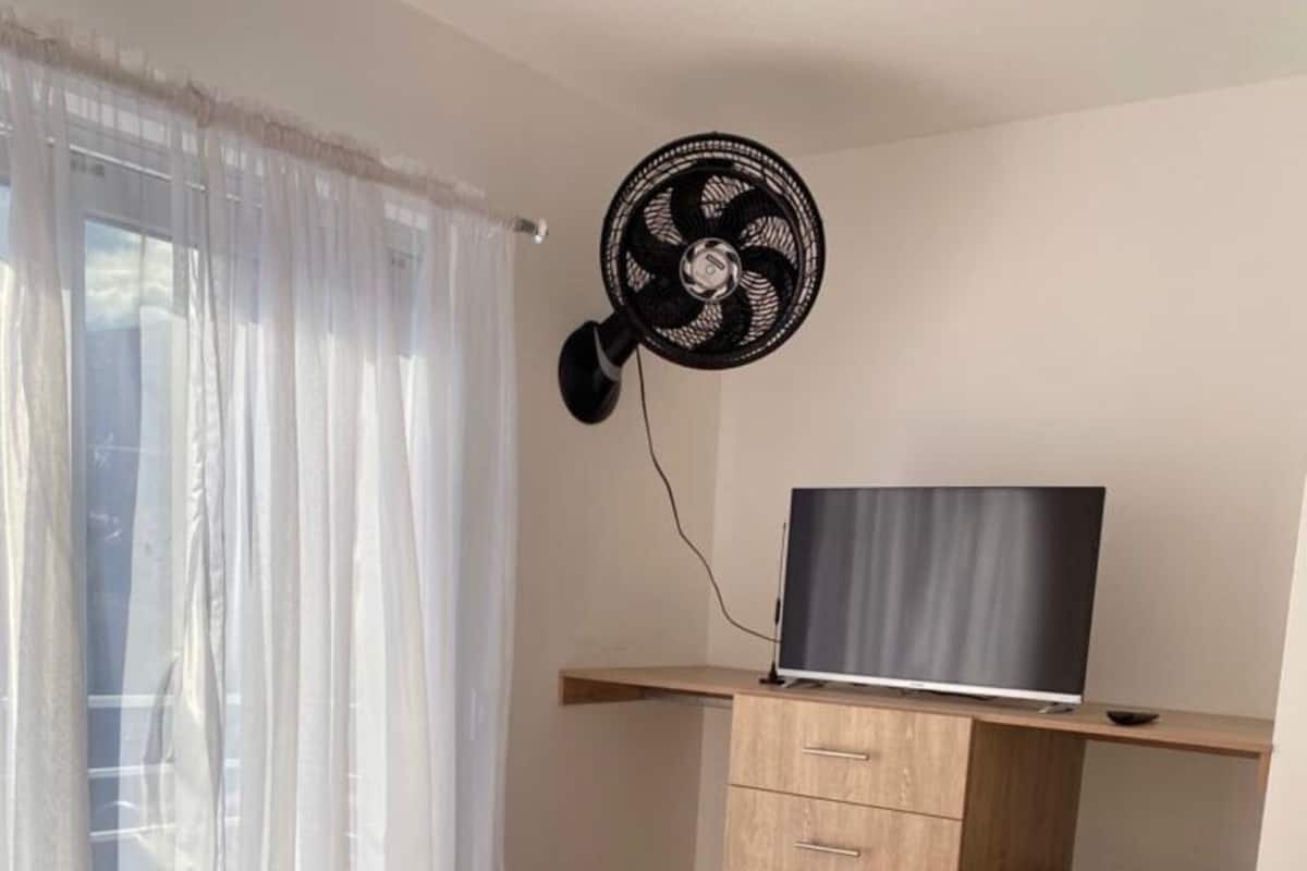 A bedroom with a bed, a TV on a wooden shelf, a fan, and a window with curtains.