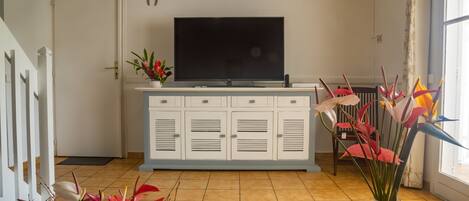 A living room with a flat-screen TV, white cabinets, a tiled floor, and a window with curtains.
