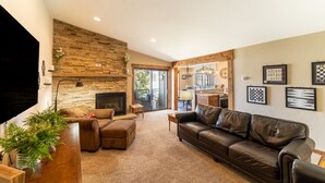A living room with a fireplace, leather sofa, and a TV. There is a kitchen area visible through an open door.