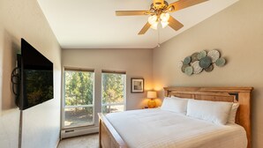 A bedroom with a large bed, a ceiling fan, a TV, and a window with a view of trees.
