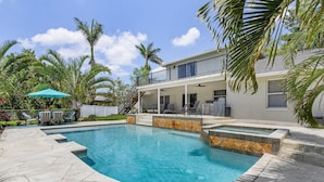 A swimming pool with a stone deck, a covered patio with chairs, and a two-story house with a balcony. - Fairview Getaway (Fort Myers)