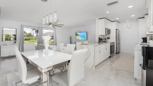 A modern kitchen with white cabinetry, a dining area with a white table and chairs, and a large window with a view of greenery. - Fairview Getaway (Fort Myers)