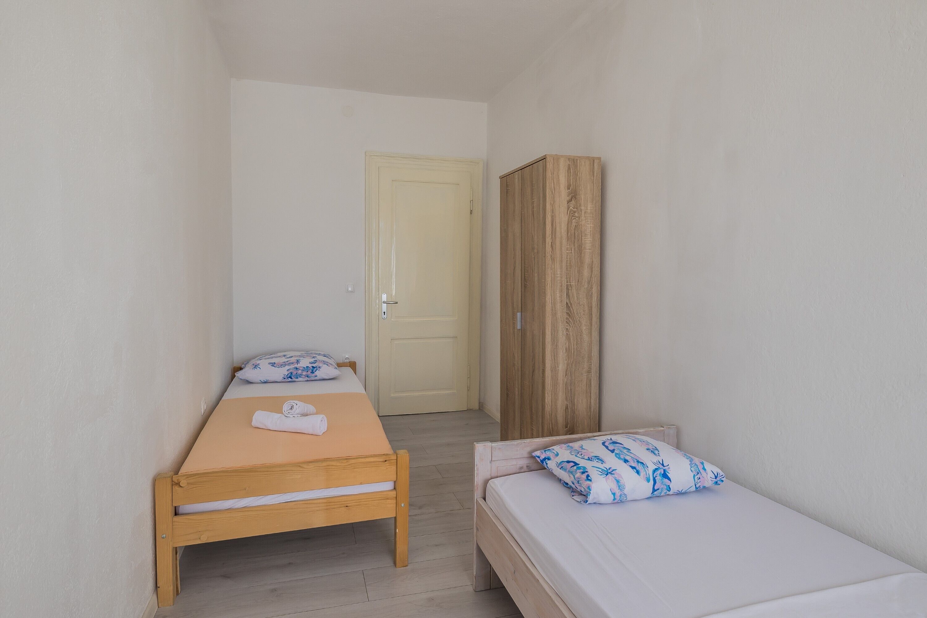 A small, narrow room with two single beds, a wooden wardrobe, and a door leading to another room.