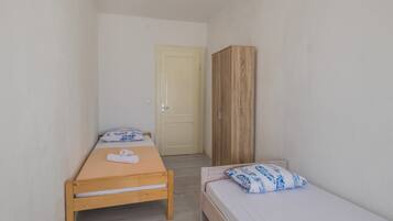 A small, narrow room with two single beds, a wooden wardrobe, and a door leading to another room.
