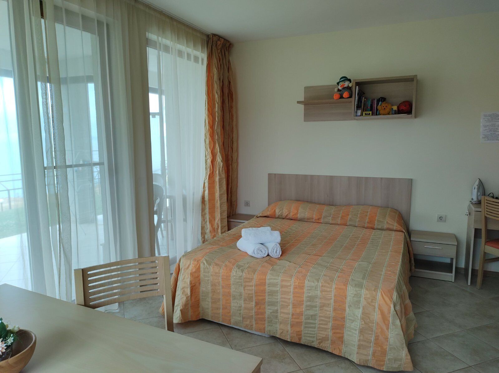 Una habitación de hotel con una cama, una mesita de noche de madera, una silla, una mesa y un estante con artículos de aseo.