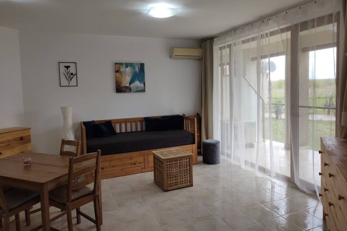 A room with a wooden sofa, a dining table with chairs, a small stool, and a trash bin. There is a window with sheer curtains and a view of the outdoors.