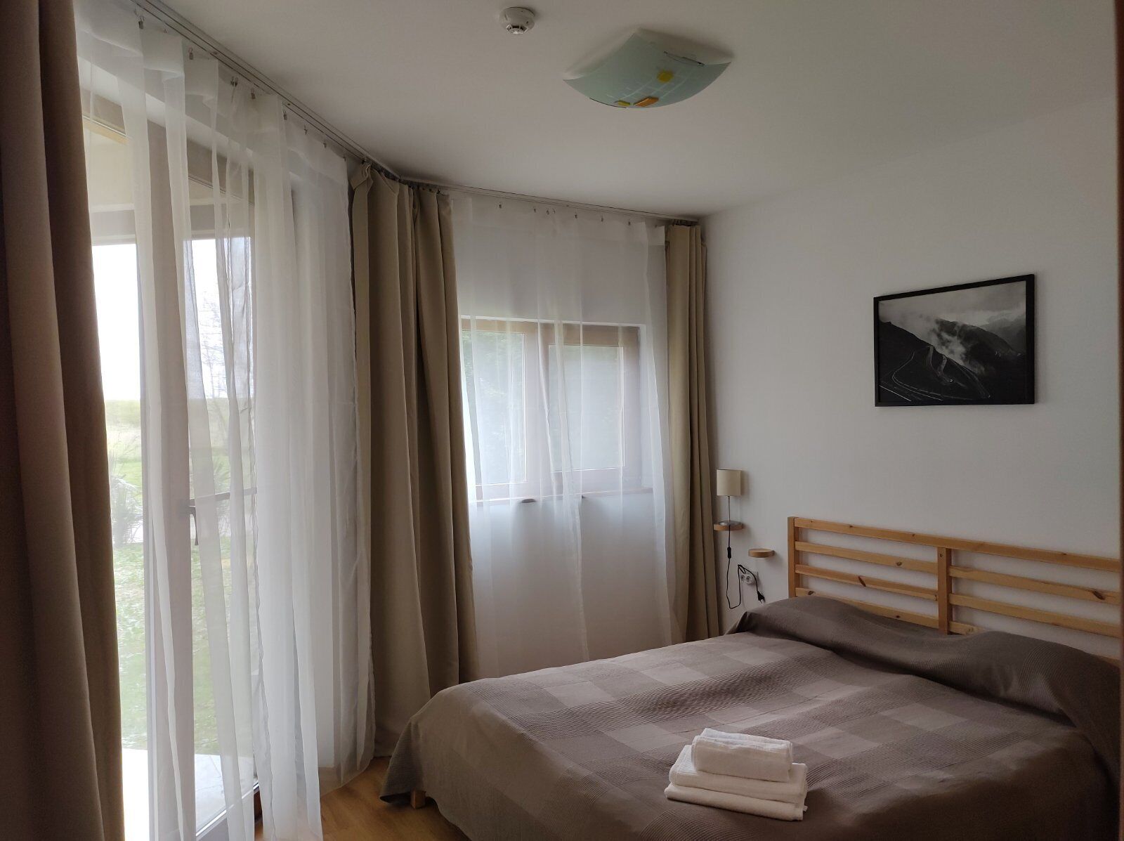 Un dormitorio con cama, cortinas, mesita de noche, lámpara y un cuadro en la pared.