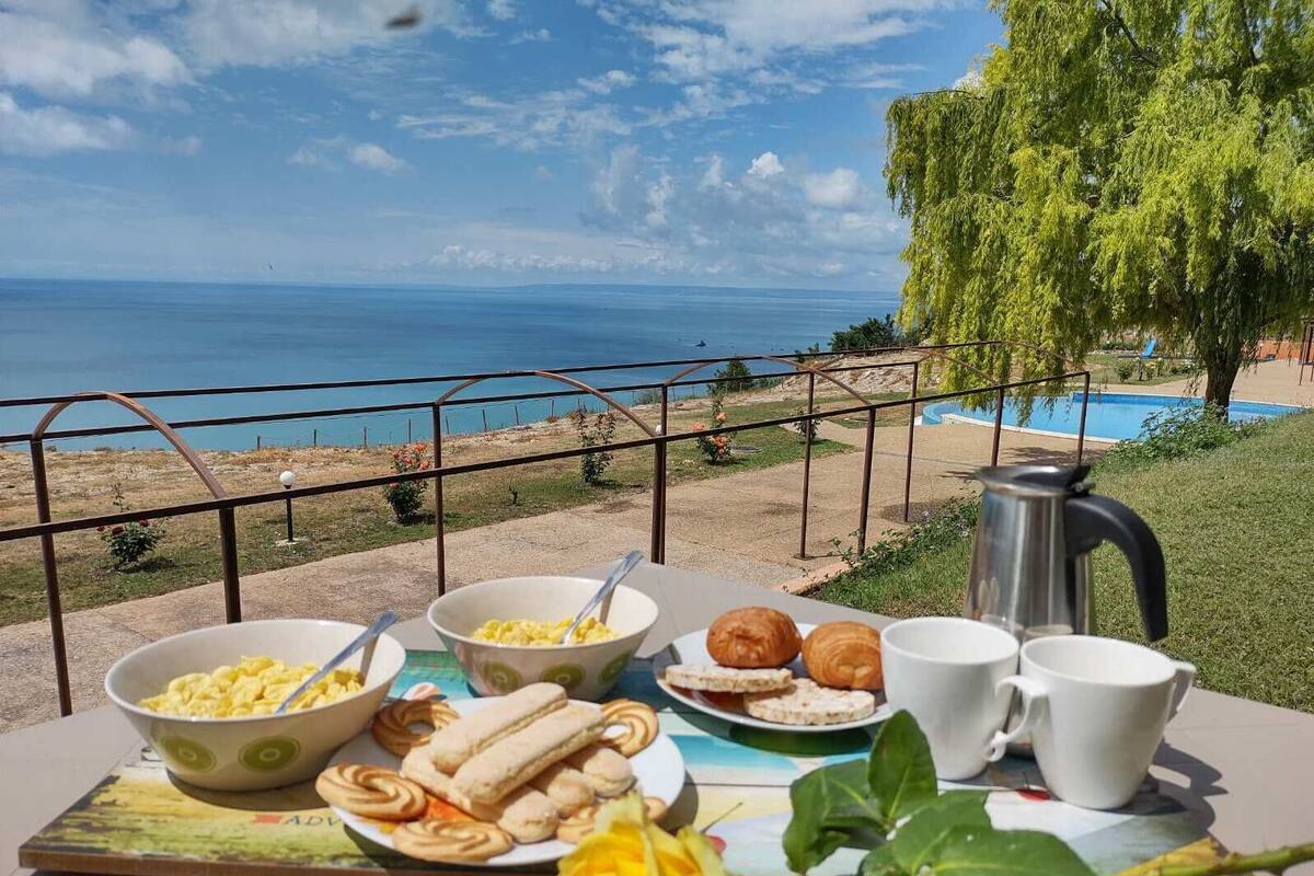 A table with breakfast items, a coffee pot, and a view of the sea and pool.