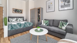 A modern living room with a grey sofa, a grey bed, and a white coffee table. There are two framed pictures on the wall, a mirror, and a wardrobe. - MARGI Maya Apartments (Szczecin)