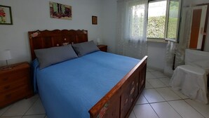 Une chambre à coucher avec un lit en bois, une table de chevet, une fenêtre avec des rideaux et une petite table accompagnée d’une chaise.