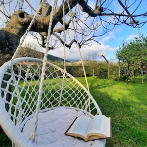 Ein weißer Korbstuhl mit Kissen, ein aufgeschlagenes Buch und eine Seilschaukel, die an einem Ast hängt.