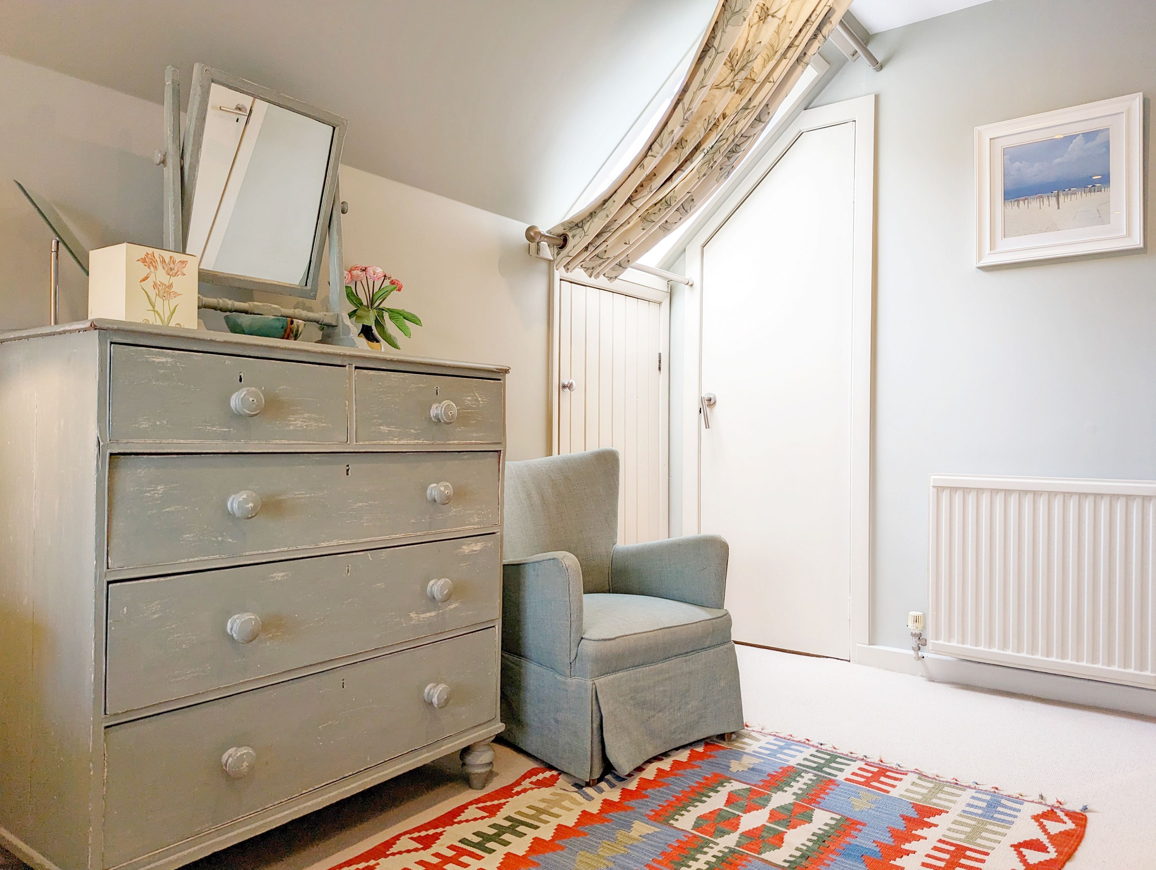 Een kamer met een grijze dressoir, een blauwe fauteuil, een kamerkleed met een patroon, een witte radiator en een ingelijst kunstwerk aan de muur.